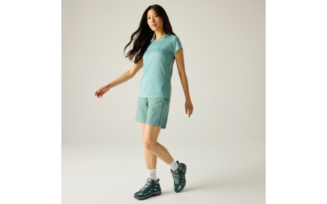 Persoon met lang donker haar, turquoise T‑shirt en shorts, witte sportsokken en groene wandelschoenen, lichte achtergrond, één voet iets opgetild