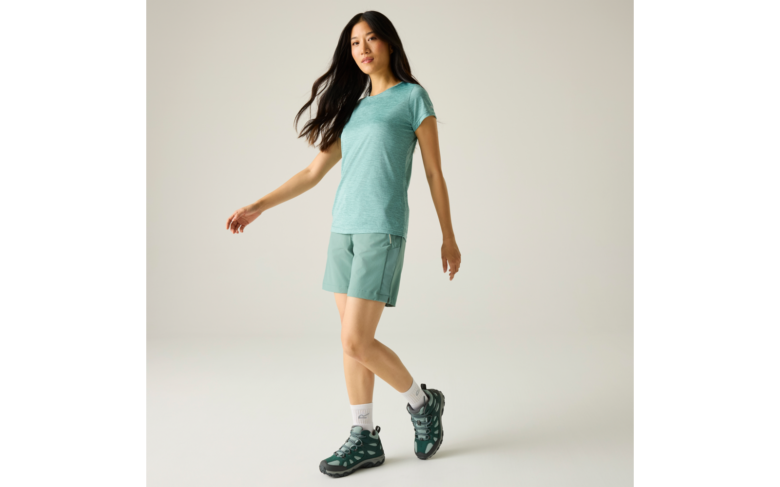 Persoon met lang donker haar, turquoise T‑shirt en shorts, witte sportsokken en groene wandelschoenen, lichte achtergrond, één voet iets opgetild