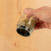 Hand hält ein Gewürzglas und streut getrocknete Kräuter auf eine Oberfläche aus Holz