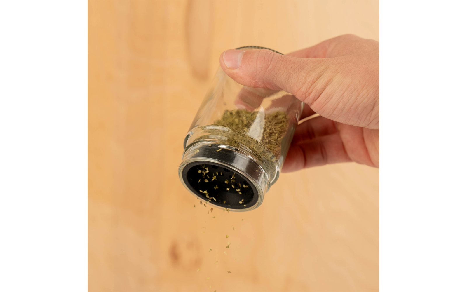 Hand hält ein Gewürzglas und streut getrocknete Kräuter auf eine Oberfläche aus Holz