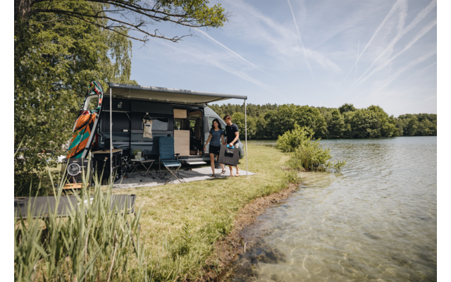 Wohnmobil mit ausgefahrener Markise am Ufer eines Sees; zwei Personen am Einstieg, befestigte Boards am Fahrzeug, Klappstühle, klares Uferwasser