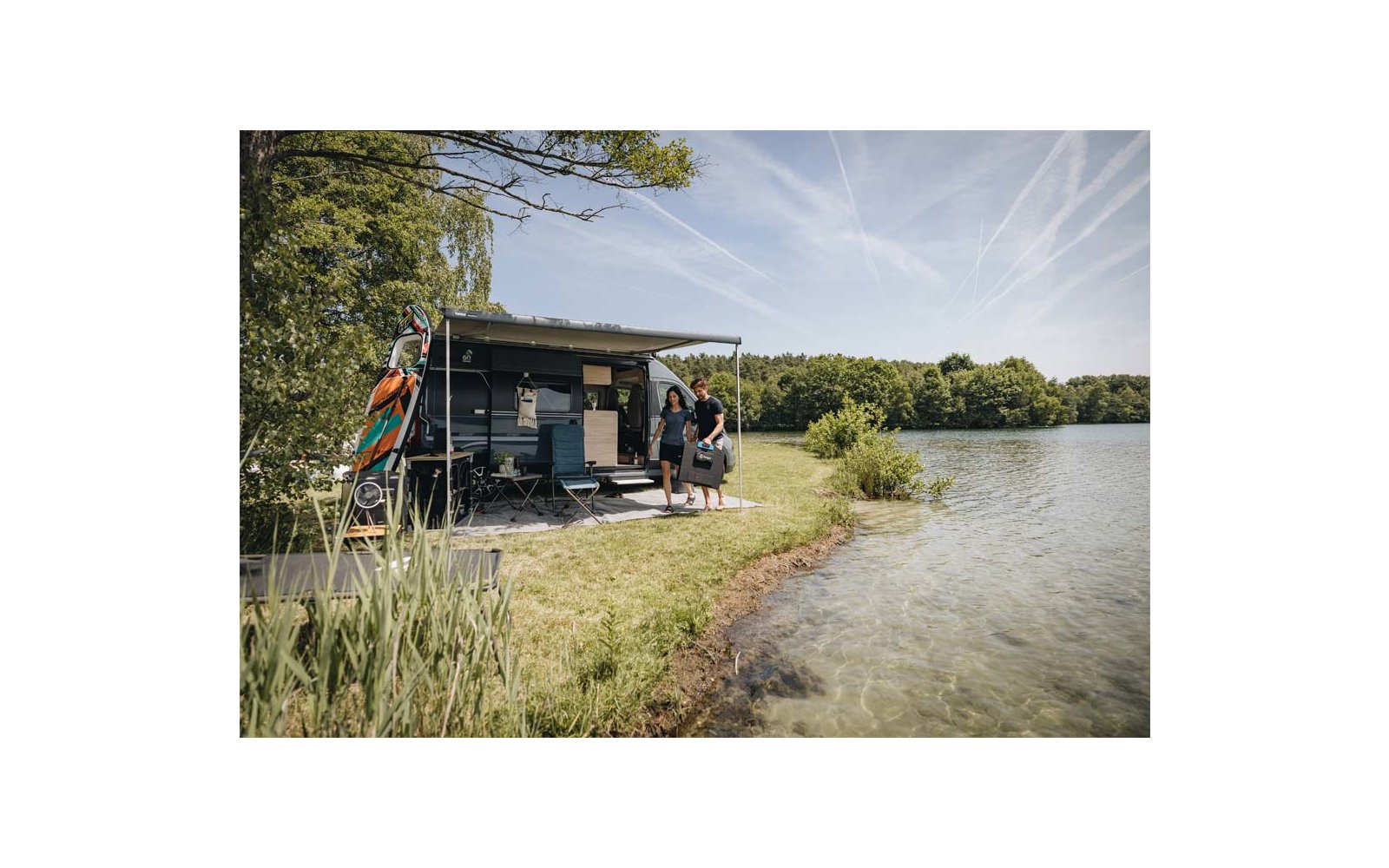 Camper con tendalino esteso sulla riva di un lago; due persone vicino all'ingresso, tavole fissate al veicolo, sedie pieghevoli e acqua limpida