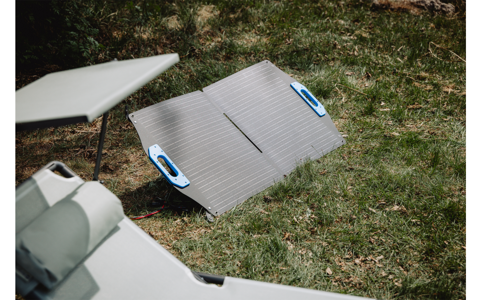 Folded rectangular panel with parallel lines and two blue handles lying on green grass; left foreground shows a gray lounge cushion