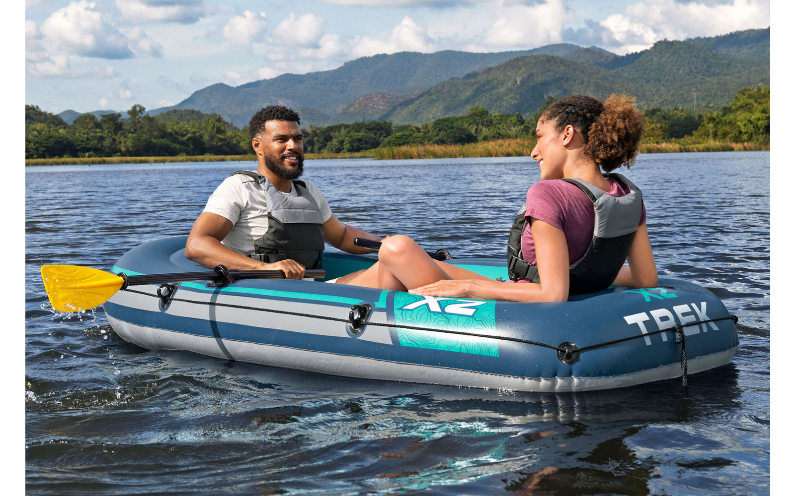 Zwei Personen sitzen in einem aufblasbaren Boot auf einem See, mit Schwimmwesten; Berge und teilweise bewölkter Himmel im Hintergrund