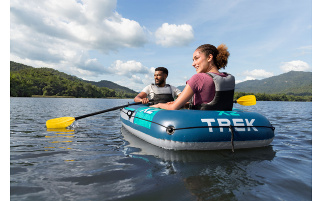 Zwei Personen in einem blauen Schlauchboot mit Aufschrift TREK, mit Schwimmwesten und gelben Paddeln, auf einem See vor bewaldeten Hügeln und wolkigem Himmel