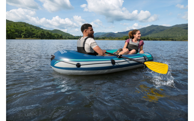 Zwei Personen in einem aufblasbaren Boot auf einem See, eine bedient ein gelbes Paddel mit Wasserspritzern; bewaldetes Ufer, Berge und wolkiger Himmel im Hintergrund