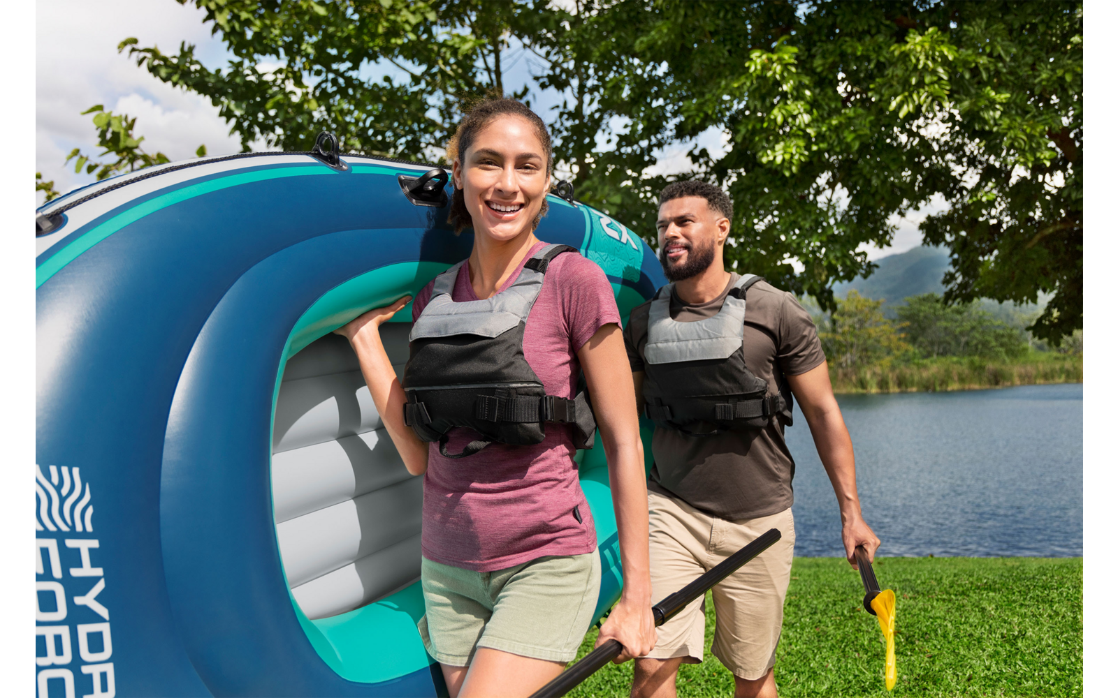 Zwei Personen mit Schwimmwesten tragen ein blau-türkisfarbenes aufblasbares Boot; eine Person hält ein Paddel, sie gehen auf Gras am Ufer eines Sees, im Hintergrund Bäume