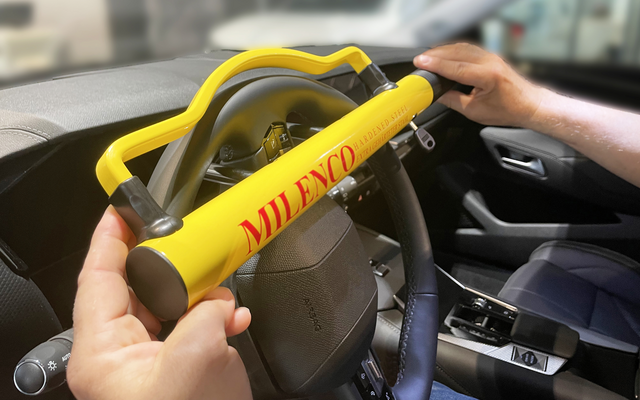 Lenkrad mit gelbem Bügel mit roter Aufschrift "MILENCO", von zwei Händen gehalten; unscharfer Auto-Innenraum im Hintergrund