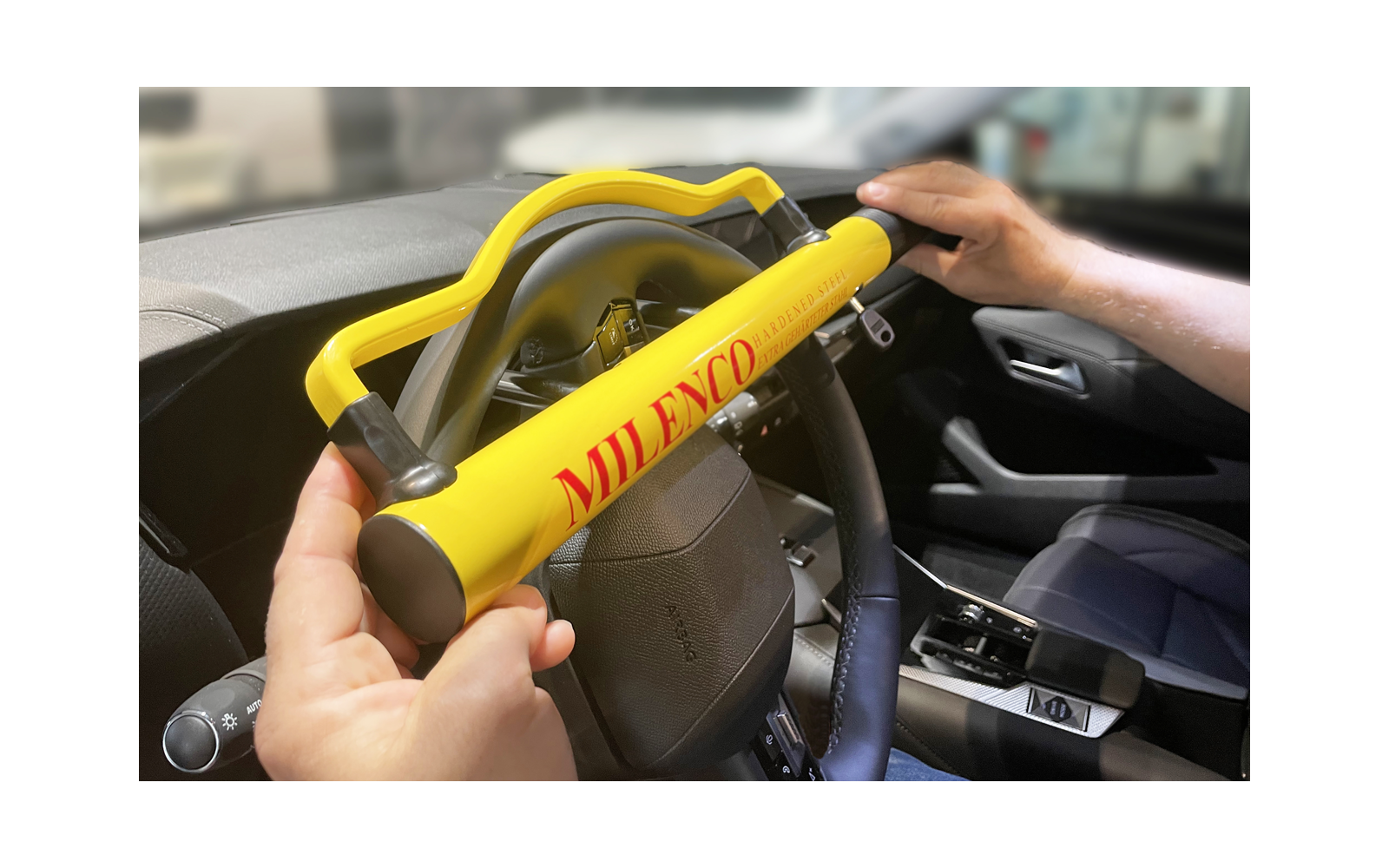 Lenkrad mit gelbem Bügel mit roter Aufschrift "MILENCO", von zwei Händen gehalten; unscharfer Auto-Innenraum im Hintergrund