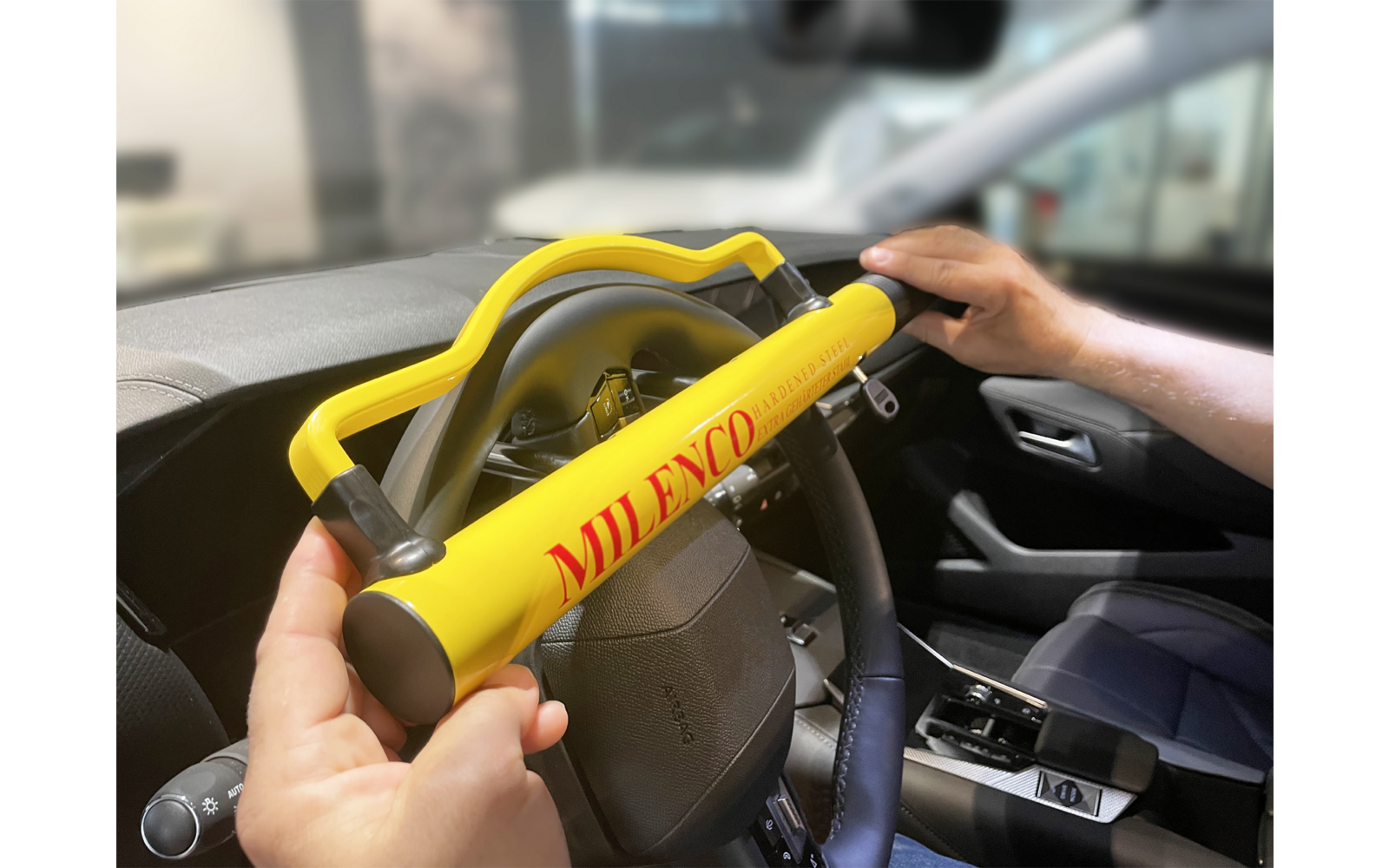 Volante con una barra gialla recante la scritta rossa "MILENCO", tenuta da due mani; interno dell'auto sfocato sullo sfondo