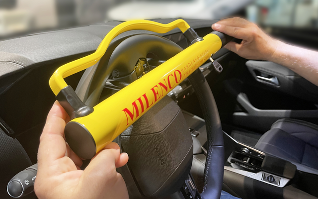 Steering wheel with a yellow bar bearing red "MILENCO" lettering, held by two hands; blurred car interior in the background