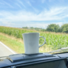 White cup placed on a dashboard platform inside a vehicle, background shows a field of green corn and a blue sky