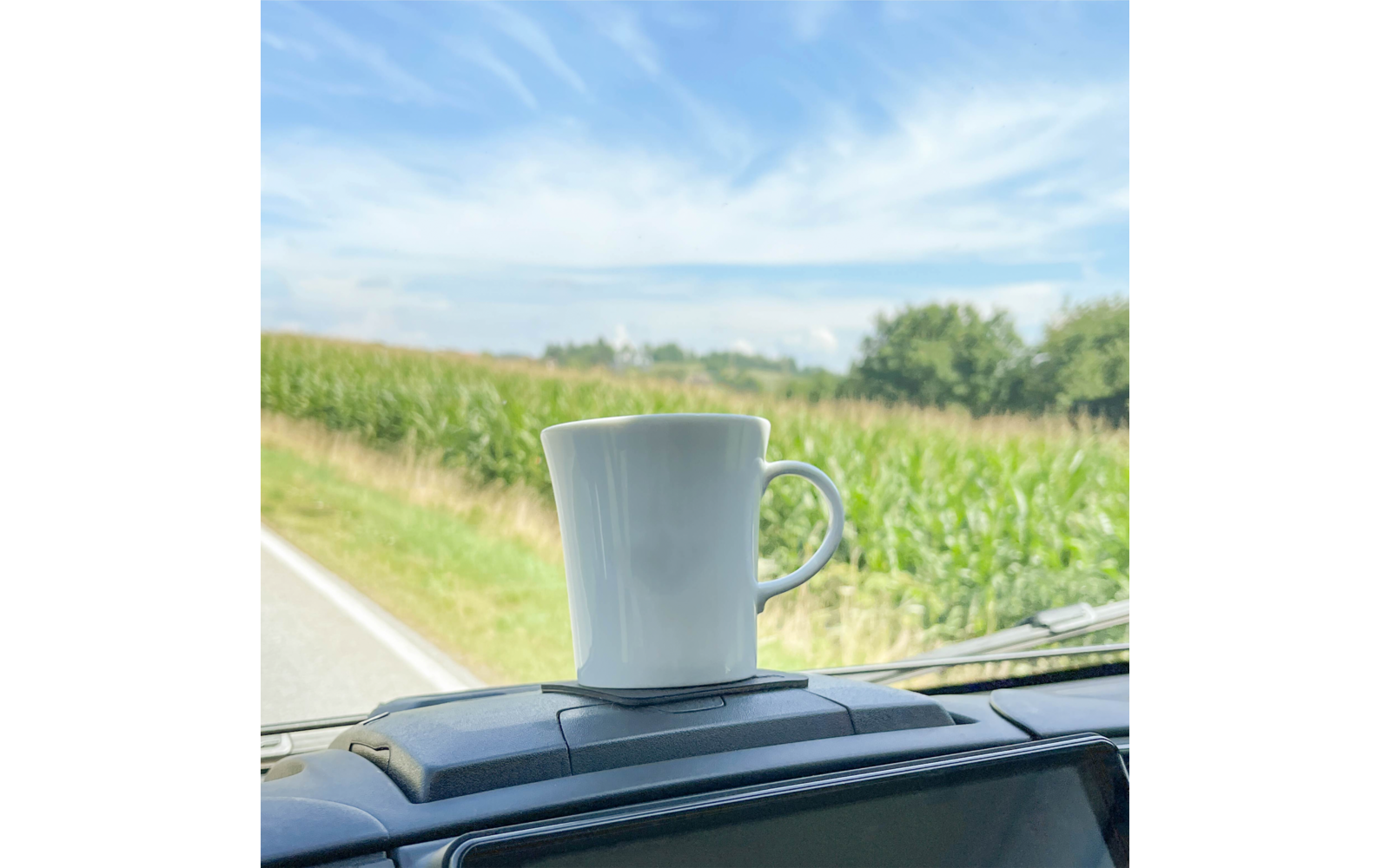 White cup placed on a dashboard platform inside a vehicle, background shows a field of green corn and a blue sky