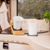 Two white mugs on a kitchen countertop, one on a wooden holder, window with trees in the background