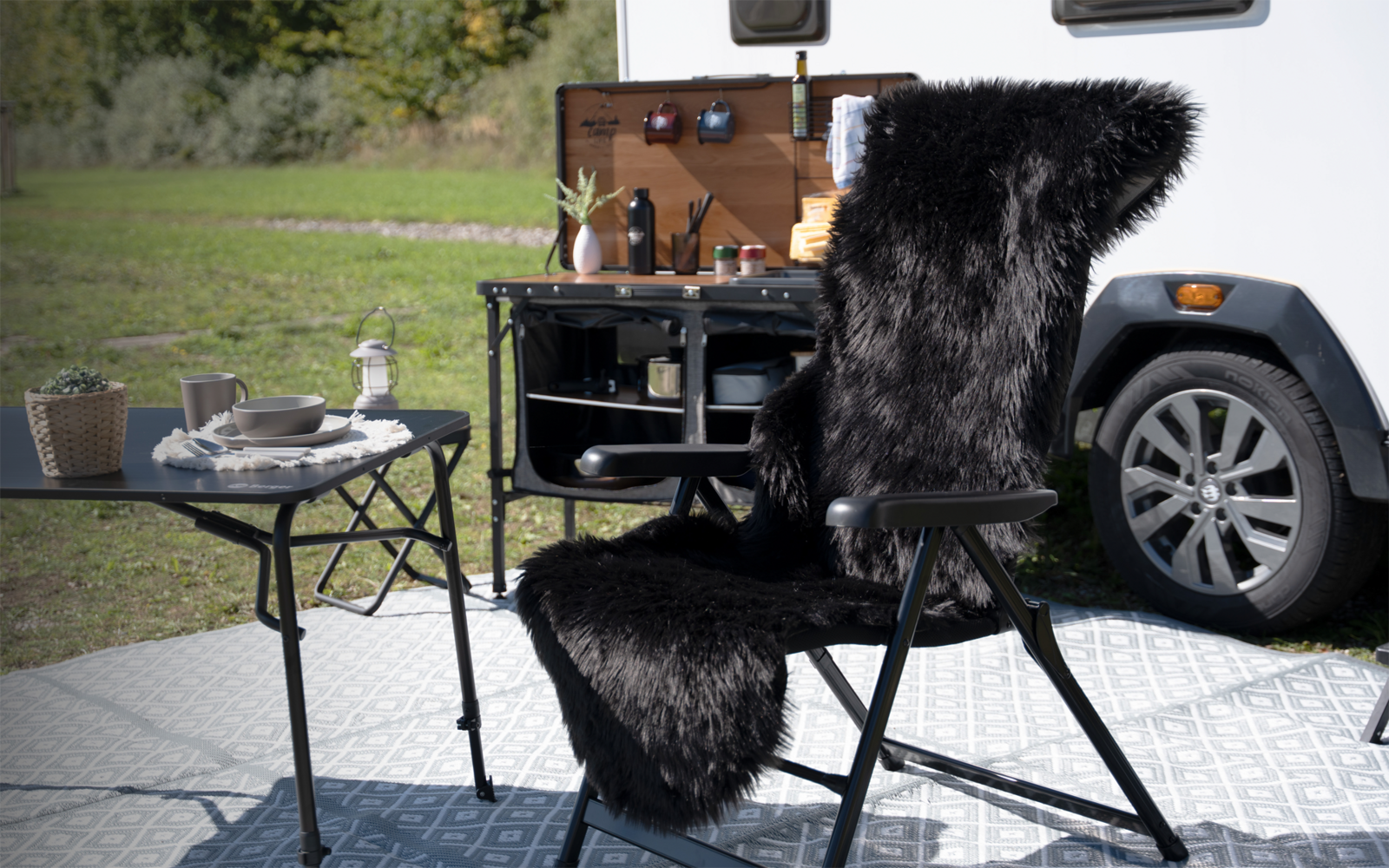Klappstuhl mit schwarzer Fellauflage, daneben klappbarer Tisch mit Tellern, Besteck und Wasserflasche; rechts Rad und Seitenwand eines Wohnwagens; im Hintergrund Grasfläche