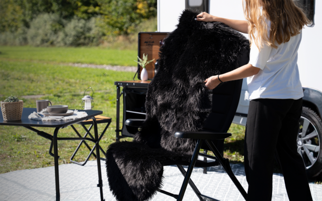 Person mit langen Haaren legt eine schwarze Fellauflage auf einen schwarzen Klappstuhl neben einem kleinen Tisch mit Teller und Tasse; Rasen und Wohnwagen im Hintergrund