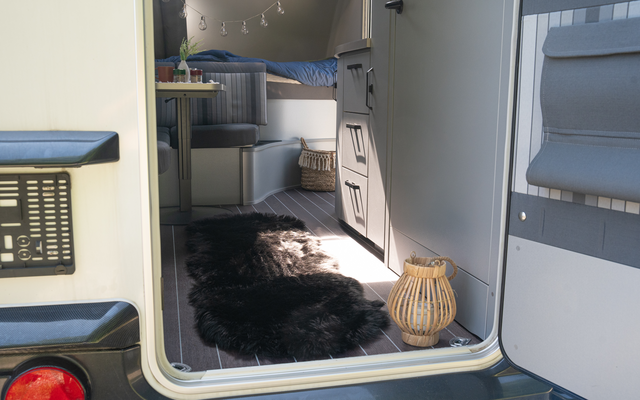 Open door of a motorhome, view of a black fur rug, woven lantern on the floor, drawers, seat cushion and a bed with blue blanket