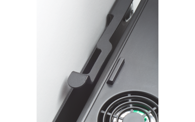 Nahaufnahme eines schwarzen Kunststoffrahmens mit hakenförmigem Element links, einem schmalen rechteckigen Knopf und einem runden Lüftergitter mit sichtbaren Flügeln und grünem Zentrum vor hellem Hintergrund