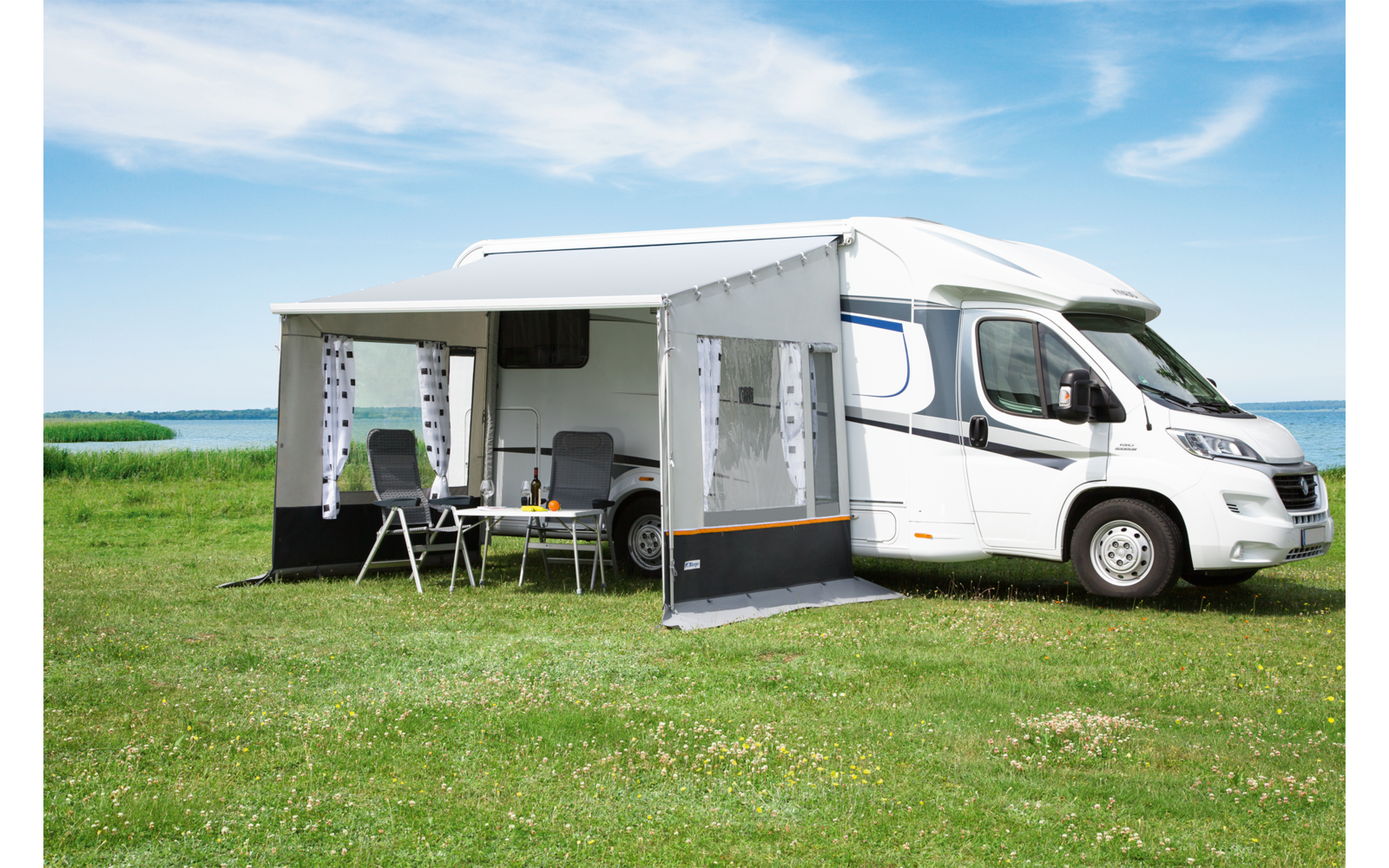 Weißes Wohnmobil mit ausgefahrener Markise und Seitenwänden, kleiner Tisch und Stühle darunter, auf grüner Wiese am Wasser; blauer Himmel mit Wolken