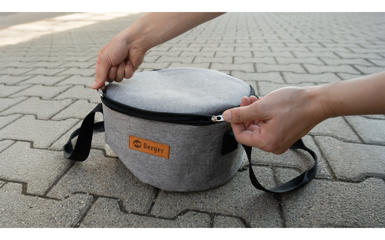 Des mains ouvrent une pochette ronde grise sur un sol pavé