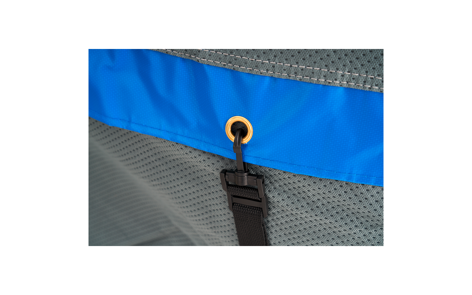Close-up of a blue fabric band with a gold metal grommet, a black cord threaded through the grommet, a black plastic buckle on a black strap, and gray mesh fabric in the background