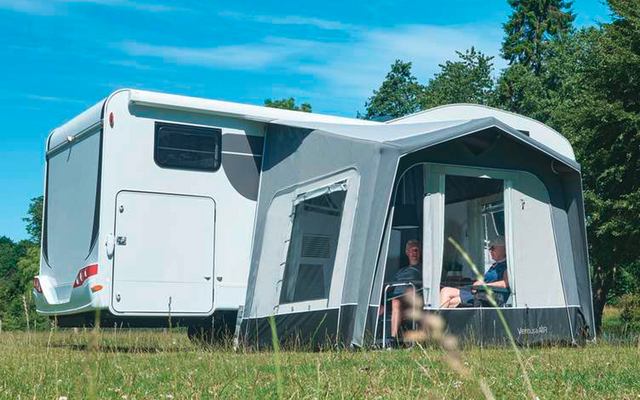 Camping-car blanc avec auvent fixé sur une prairie, arbres en arrière-plan et ciel bleu