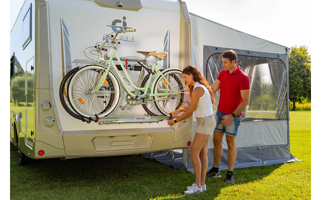 Frau und Mann an der Rückseite eines Wohnmobils, zwei Fahrräder auf einem Heckträger, graue Markise ausgefahren, Wohnmobil auf einer Wiese