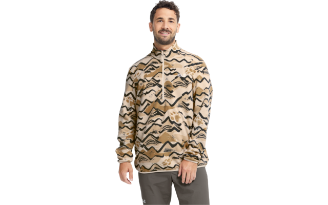 Person steht, trägt beige Zip‑Pullover mit schwarzen, wellenartigen Mustern und dunkelgraue Hose