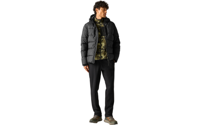 Persoon met krullend haar draagt een zwarte gewatteerde jas, camouflage-hoodie, zwarte joggingbroek en groene sneakers, staat op een lichte achtergrond met handen in de zakken, kijkt naar links