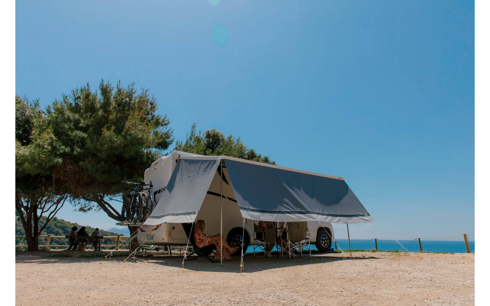 Wohnwagen mit ausgeklapptem Vordach auf trockenem, kiesigem Boden, Klappstühle und Tisch unter dem Vordach, links zwei Personen an einem Picknicktisch unter einem Baum, Bäume, blauer Himmel und Meer am Horizont
