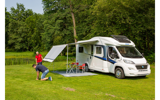Witte camper op gras met uitgeklapte luifel, twee personen die een grondzeil uitrollen; twee rode klapstoelen, klein tafeltje, bomen en een vijver op de achtergrond