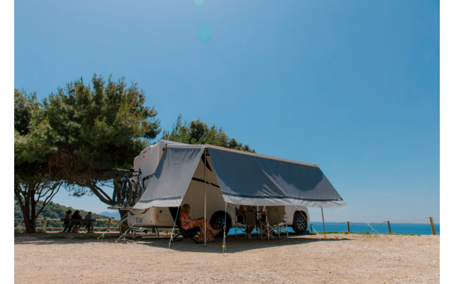 Wohnwagen mit ausgeklapptem Vordach auf trockenem, kiesigem Boden, Klappstühle und Tisch unter dem Vordach, links zwei Personen an einem Picknicktisch unter einem Baum, Bäume, blauer Himmel und Meer am Horizont