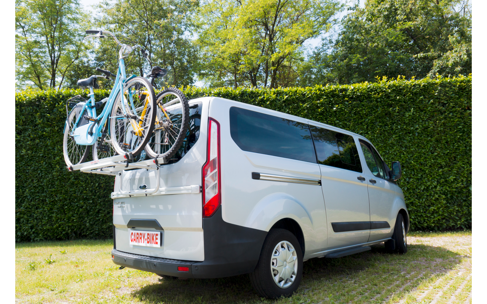 Weißer Transporter mit zwei Fahrrädern auf einem Heck-Fahrradträger, geparkt auf Gras vor einer Hecke und Bäumen an einem sonnigen Tag