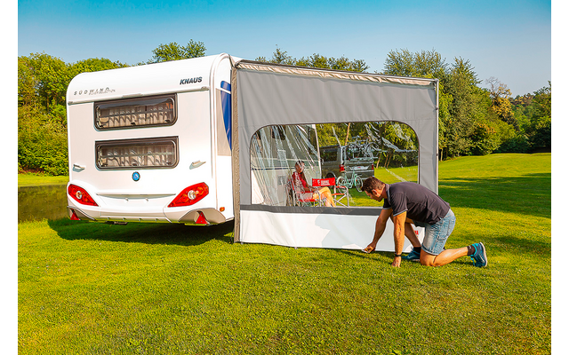 Weißer Wohnwagen auf grüner Wiese mit seitlich befestigtem Vorzelt und Fenster; eine Person kniet am Vorzelt, im Zelt Tisch, Stühle und rote Gegenstände sichtbar; Bäume und blauer Himmel im Hintergrund
