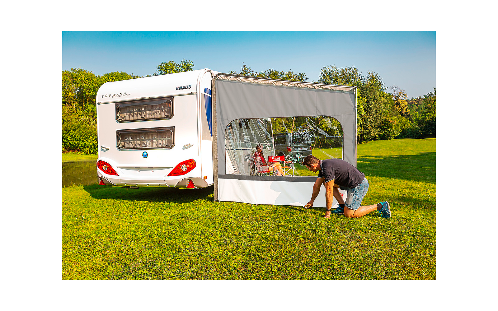Weißer Wohnwagen auf grüner Wiese mit seitlich befestigtem Vorzelt und Fenster; eine Person kniet am Vorzelt, im Zelt Tisch, Stühle und rote Gegenstände sichtbar; Bäume und blauer Himmel im Hintergrund