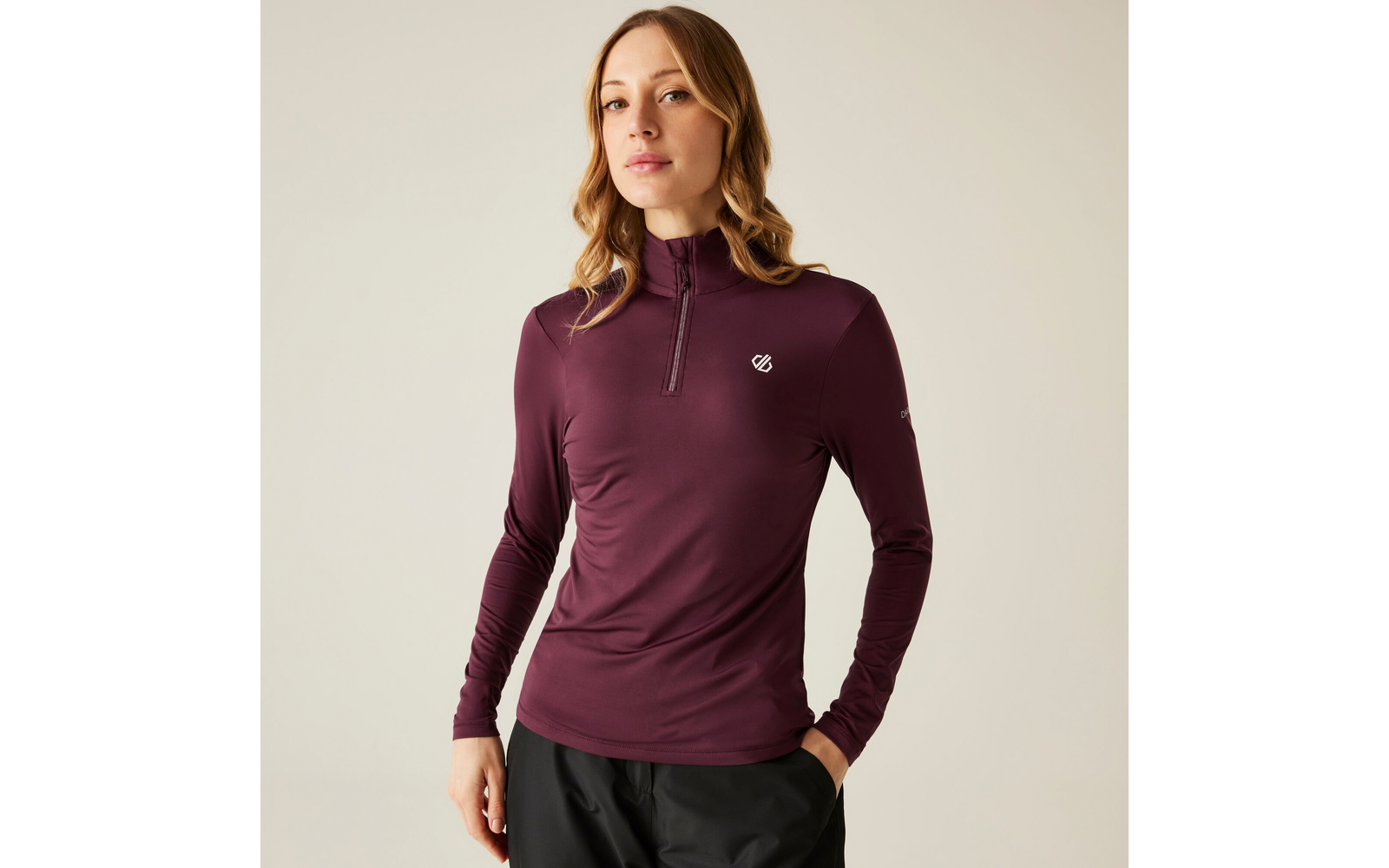 Femme aux longs cheveux bouclés porte un haut à manches longues violet foncé avec fermeture éclair et un pantalon noir sur fond neutre