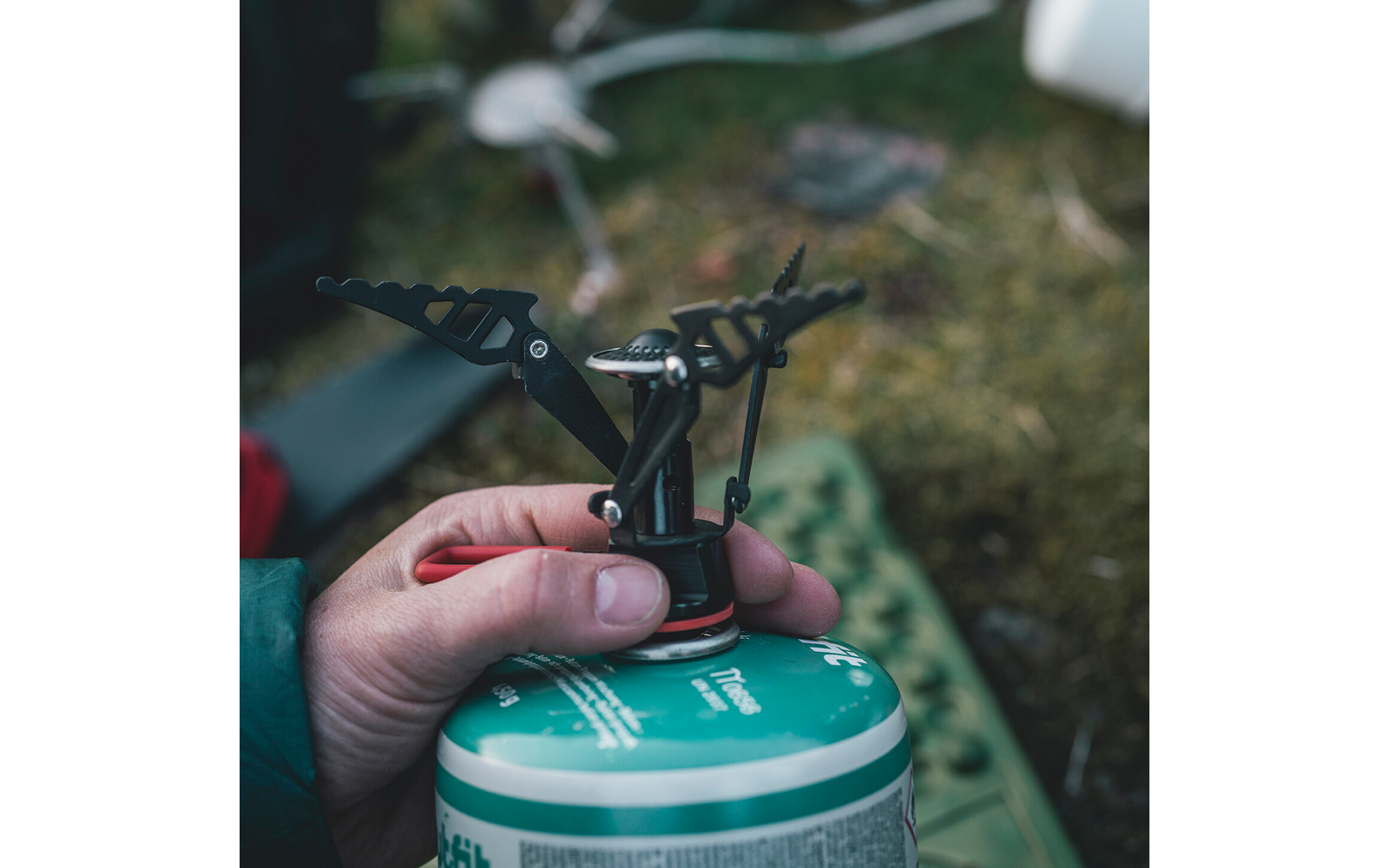 Hand houdt een kleine campingbrander op een groene gasbus, gras en onscherpe kampeerspullen op de achtergrond