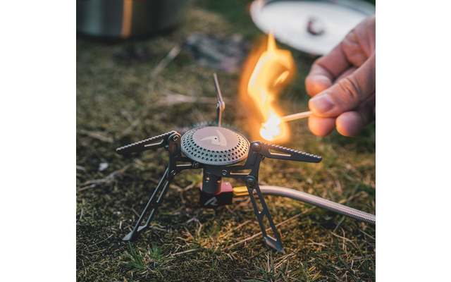 Hand steekt lucifer aan bij draagbare gasbrander met uitgeklapte poten op gras; onscherpe deksel op de achtergrond