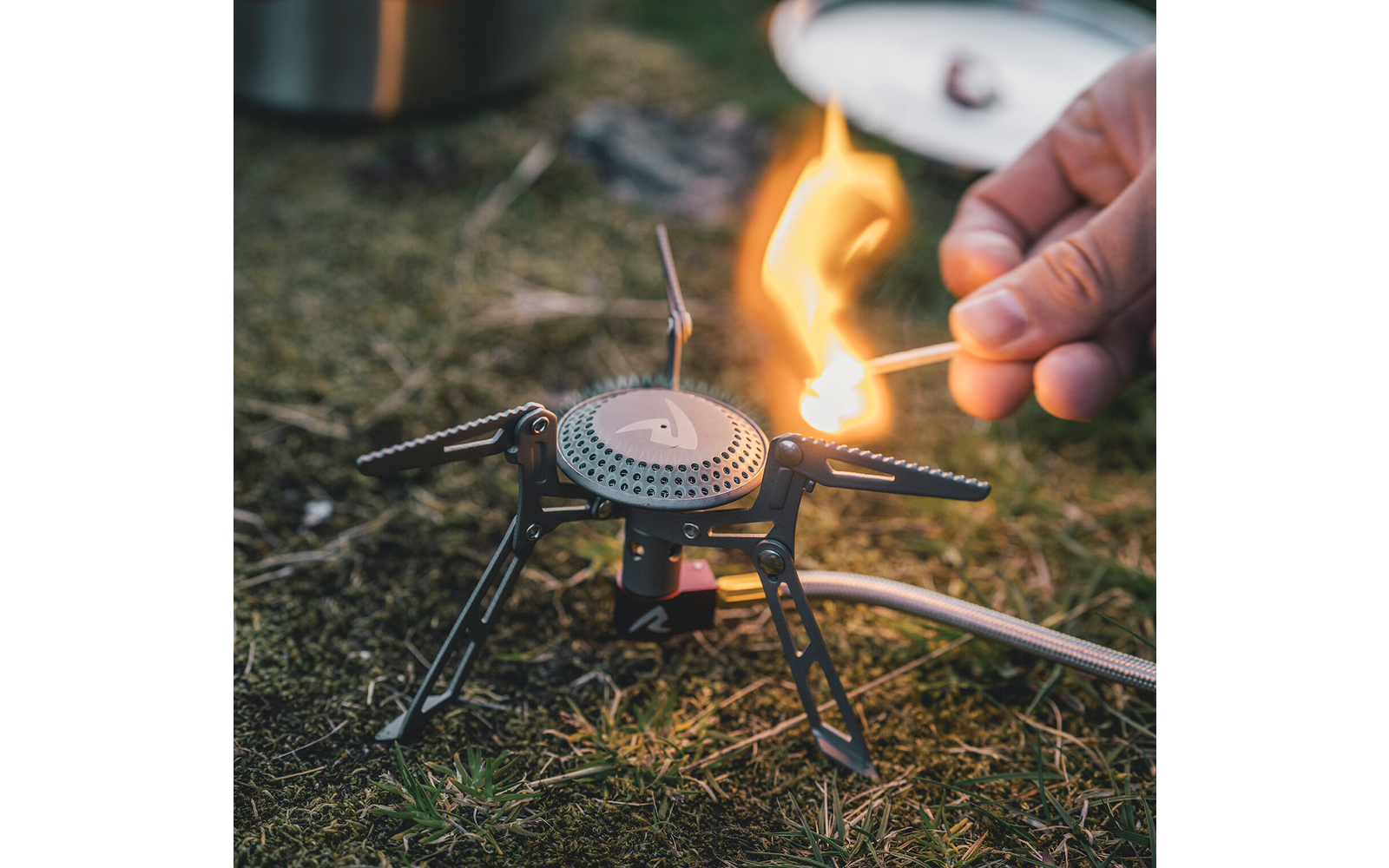 Hand steekt lucifer aan bij draagbare gasbrander met uitgeklapte poten op gras; onscherpe deksel op de achtergrond