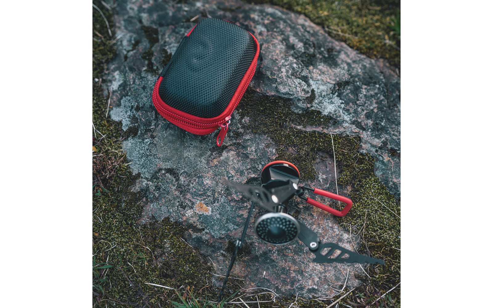 Opvouwbare gasbrander met rode handgrepen naast een zwarte harde etui met rode rits op een mosbedekte rots