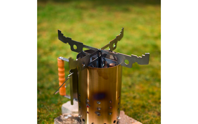 Zylindrischer goldfarbener Metallbehälter mit Belüftungslöchern, oben vier zusammengeklappte schwarze Metallträger, seitlicher Holzgriff, auf Steinen auf grünem Rasen