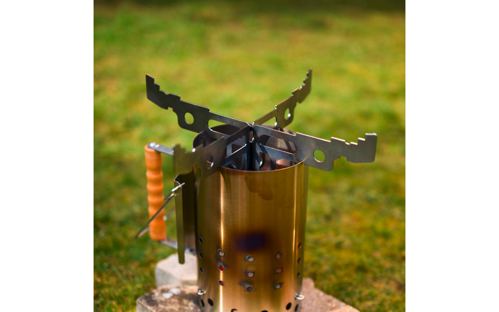 Zylindrischer goldfarbener Metallbehälter mit Belüftungslöchern, oben vier zusammengeklappte schwarze Metallträger, seitlicher Holzgriff, auf Steinen auf grünem Rasen