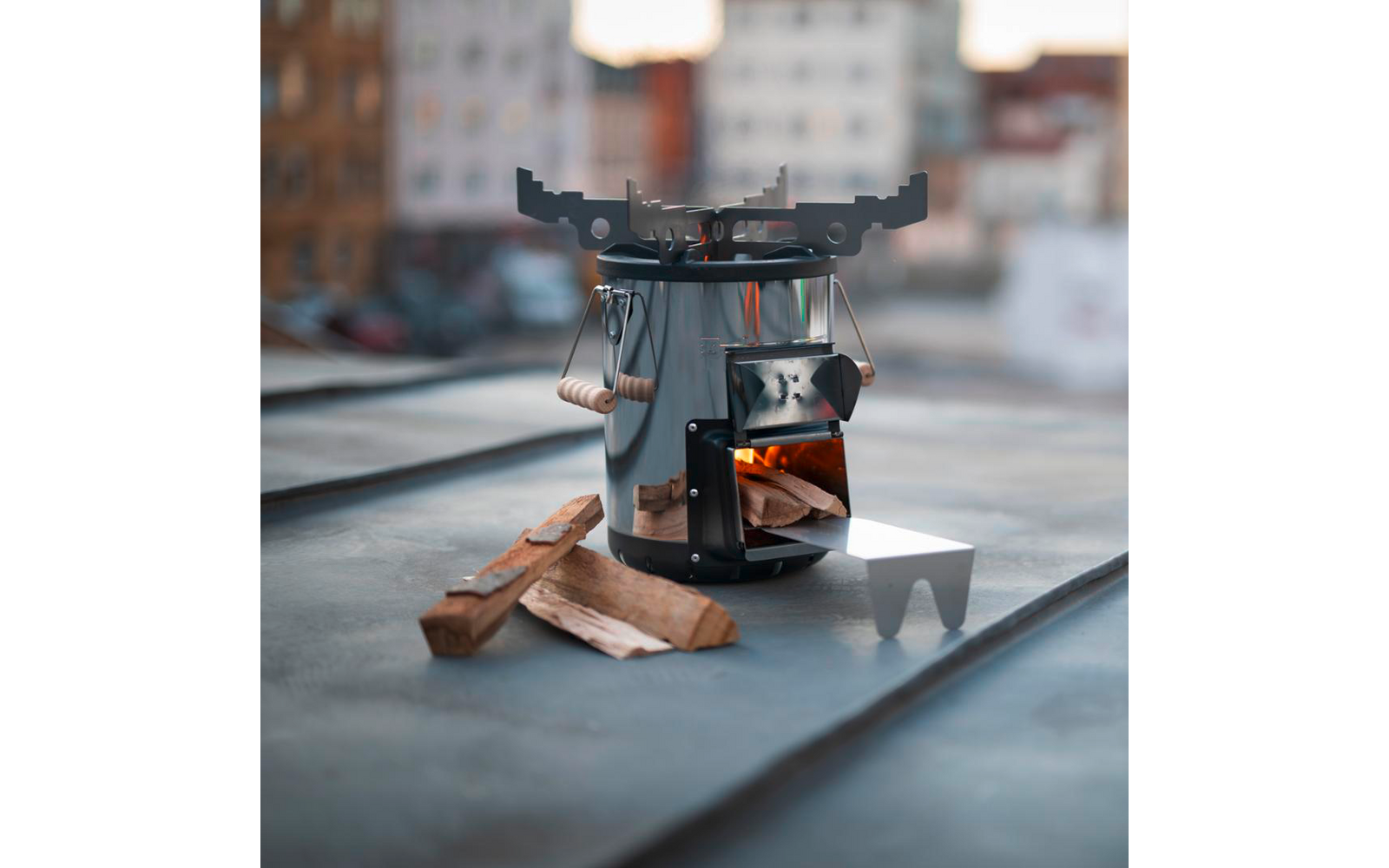 Metallischer zylindrischer Holzofen mit offener Tür und brennendem Holz, Topfhalter oben, Holzscheite daneben auf einem Metall-Dach; verschwommene Gebäude im Hintergrund