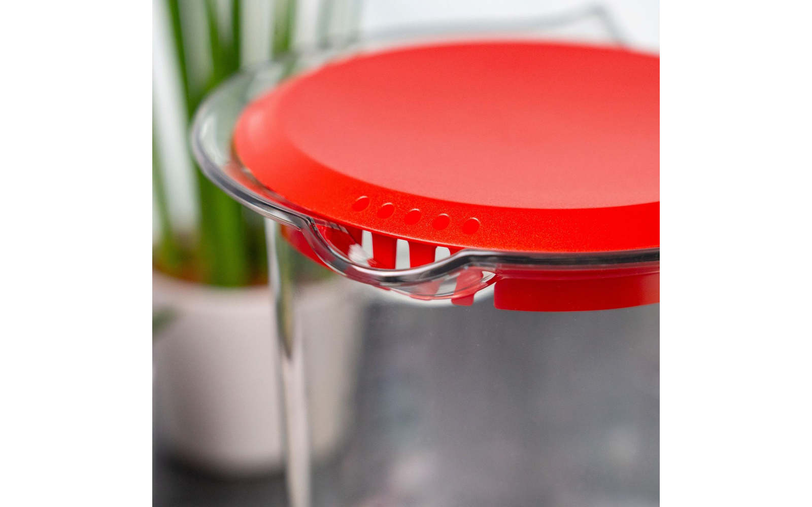Doorzichtige plastic container met rood deksel, kleine nopjes op de rand en schenktuit; onscherpe achtergrond met witte plantenpot en groene bladeren