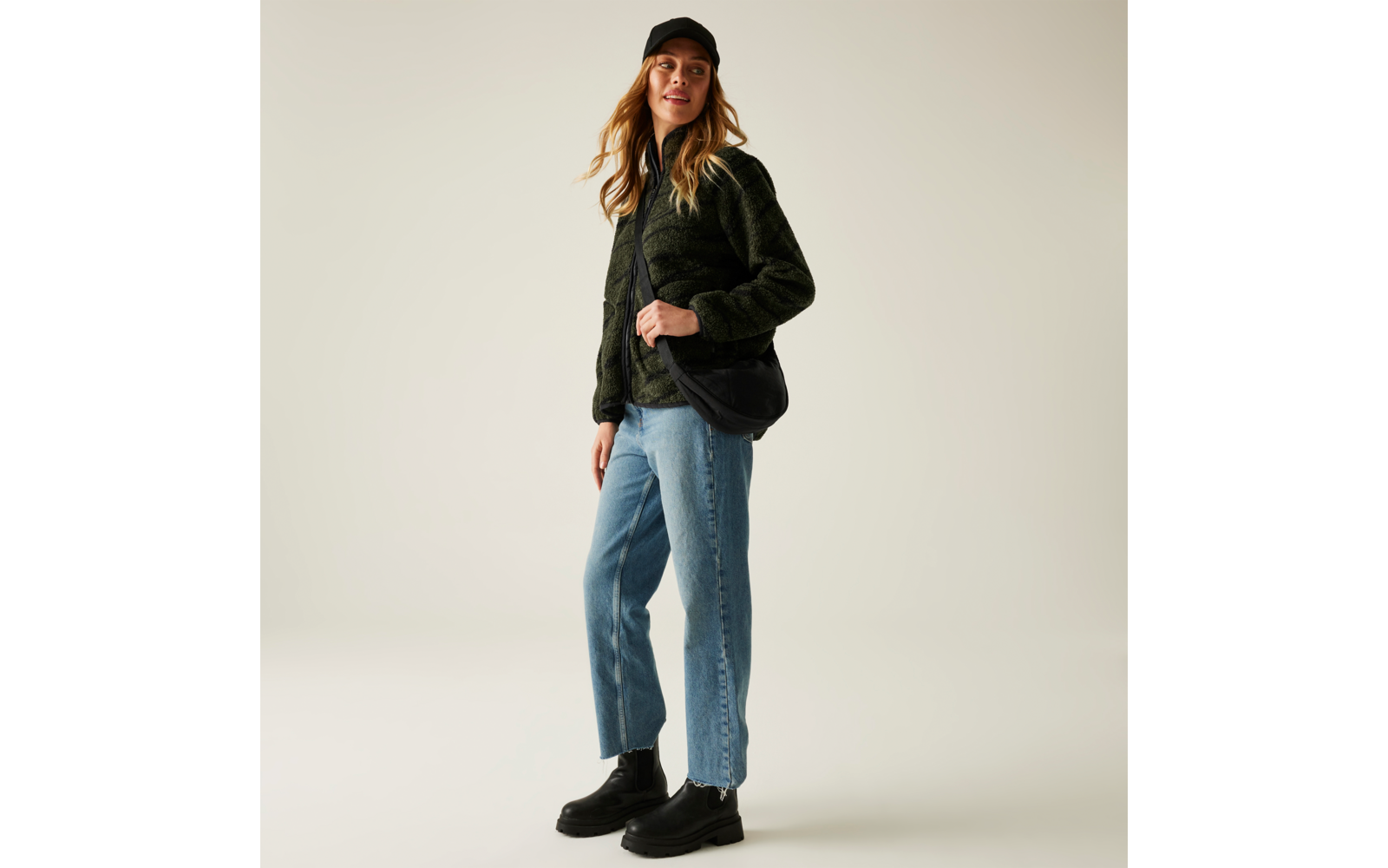 Frau steht vor hellem Hintergrund, trägt schwarze Mütze, schwarze Jacke, blauen Pullover, Jeans und schwarze Schuhe