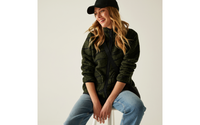 Frau mit langer blonder Haaren, schwarzem Hut, schwarzem Pullover und blauer Jeans sitzt lächelnd auf einem Hocker