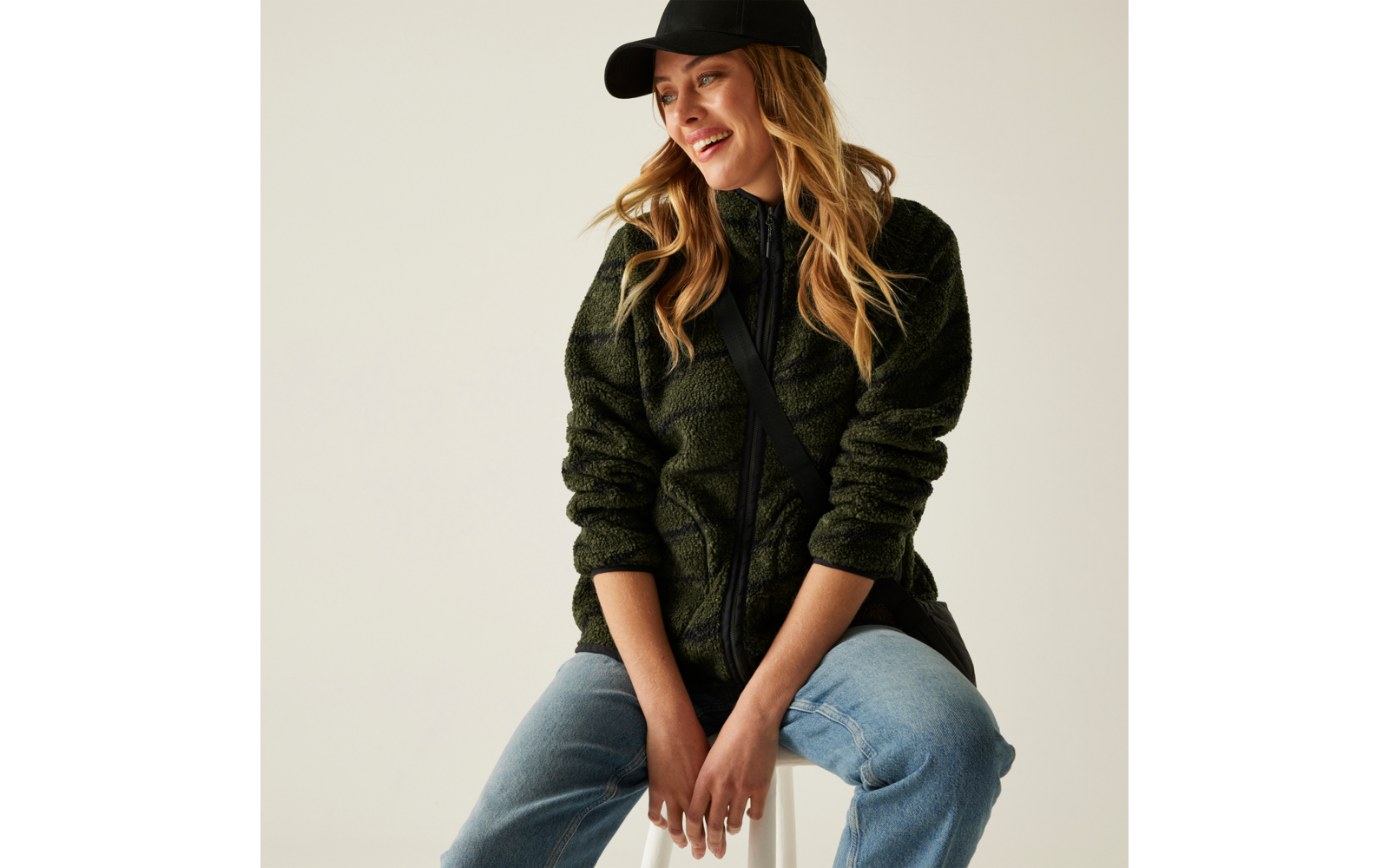 Frau mit langer blonder Haaren, schwarzem Hut, schwarzem Pullover und blauer Jeans sitzt lächelnd auf einem Hocker