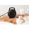 Zwei Hände halten weißen Stoff unter einer Nähmaschine; links auf dem Holztisch eine schwarze Kabeltrommel, orange Schere, Stecknadelbox und Garn