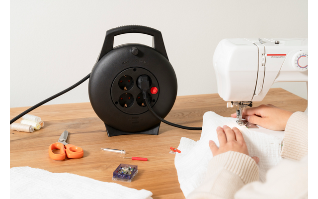 Zwei Hände halten weißen Stoff unter einer Nähmaschine; links auf dem Holztisch eine schwarze Kabeltrommel, orange Schere, Stecknadelbox und Garn