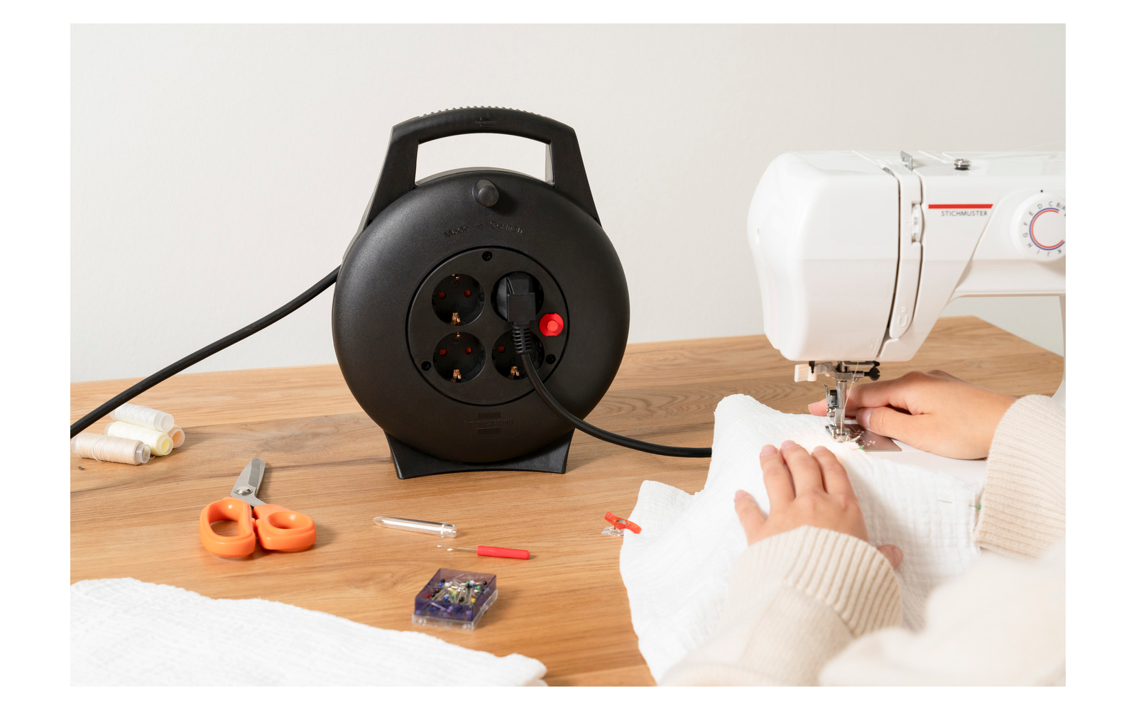 Zwei Hände halten weißen Stoff unter einer Nähmaschine; links auf dem Holztisch eine schwarze Kabeltrommel, orange Schere, Stecknadelbox und Garn